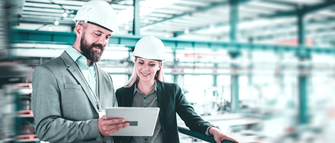 A man and a woman are standing next to each other in an industrial hall. The man holds a tablet in his hand and shows the woman the camos CPQ solution on the tablet.